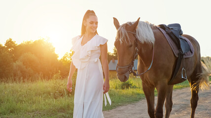 Lady brunette with long hair in ponytail wanders with horse with saddle along ground road against green meadow under bright morning sunlight