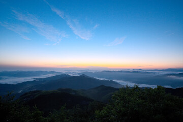 When setting sun goes down, mountain is foggy under blue and orange sky.