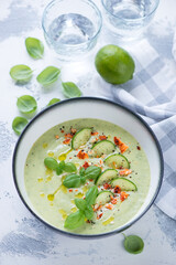 Bowl of cold cucumber gazpacho topped with cheese and green basil, vertical shot on a white concrete background