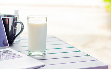 The picture of milk in a clear glass laying on a desk with a laptop is a healthy food supply in the morning to get ready for work with copy space for text.