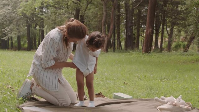 Young woman changing toddler son nappy while spending time outdoors in park on sunny day