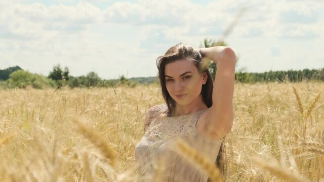 Beautiful sexy woman in field of rye or wheat, playfully looks at camera and straightens her hair. Portrait of pretty lady in dress in the middle of nowhere