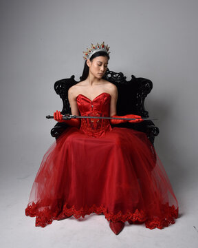 Full length  portrait of beautiful young asian woman wearing red corset, long opera gloves and ornate gothic queen crown. Graceful posing while seated on chair, isolated on studio background.