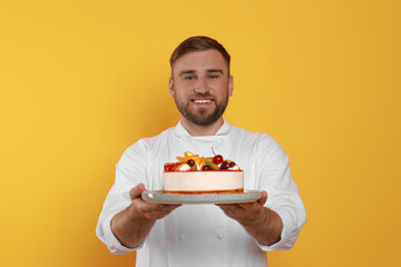 Happy professional confectioner in uniform holding delicious cake on yellow background