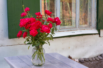 A bouquet of red roses on a table near the window of an old village house with green shutters on a summer day.
