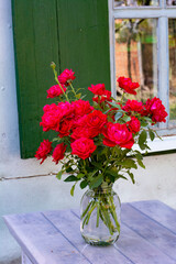 Fototapeta na wymiar A bouquet of red roses on a table near the window of an old village house with green shutters on a summer day.