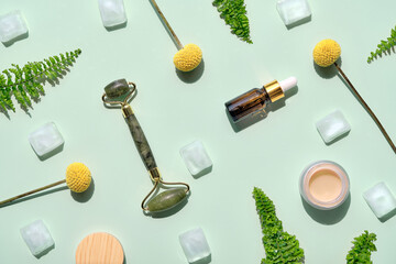 Fototapeta na wymiar Green jade face roller with ice , cream and essential oil. Mint green flat lay with exotic plants. Fern leaves, Craspedia ping pong flowers. Beauty facial with home made moisturizer. Geometric