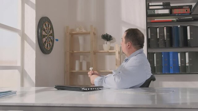 Middle aged man in blue shirt at workplace in office throwing darts at dartboard on wall during working hours.