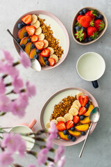 Delicious healthy breakfast, granola with yogurt and sweet fruits, banana and strawberries, blueberries and peaches, milk in a mug and flowers in a vase, top view
