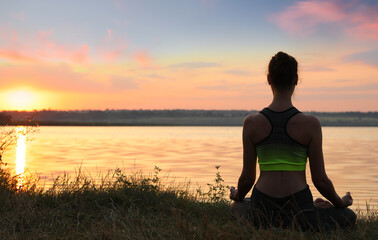 Woman meditating near river at sunset, back view. Space for text