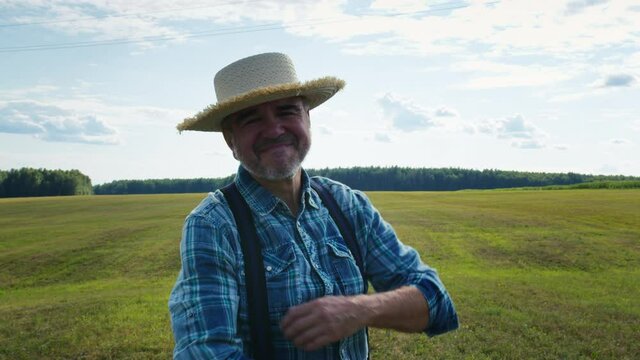 Smiling elderly farmer portrait. Agronomist wearing straw hat looking in camera with smile, standing in field plantation. Happy senior man with grey beard posing on landscape background.