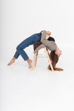 young caucasian attractive woman with long brown hair in blue jeans, suit jacket bending her body on white background. skinny female lying in bridge position on wooden ladder at studio with bare feet