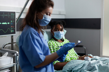 Obraz premium Nurse examining patient healthcare and illness immunity sitting in hospital ward. Caucasian woman checking treatment progress on young african american adult laying in bed at medical clinic