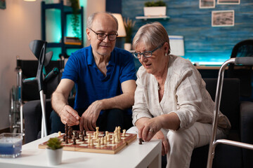 Old retired husband and wife enjoying chess game playing on board table in living room at home. Senior married couple enjoying moment on couch with crutches walk frame and wheelchair