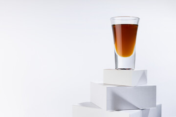 Golden scotch in shot glass in sunlight with shadows on white square podium with reflections in soft light abstract space in minimal modern style.