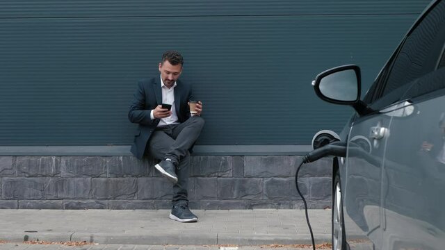 Businessman uses phone and drinks coffee while waiting for electric car to finish charging. Man in jacket resting while charged EV batteries in electric car in parking lot