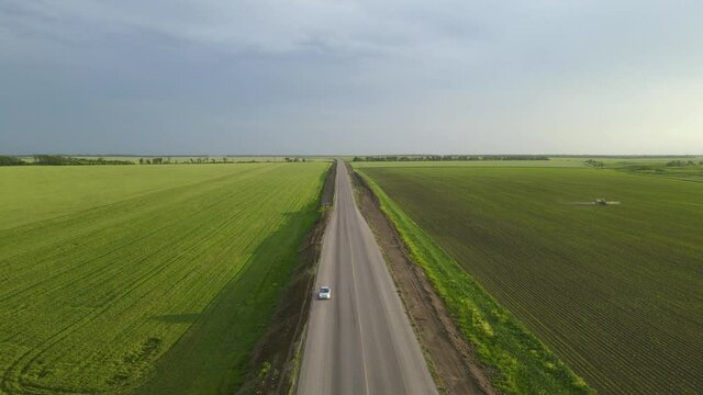 Grey asphalt road with automobile standing between green fields with tractor sprinkling fertilizers UNDER blue spring sky aerial view