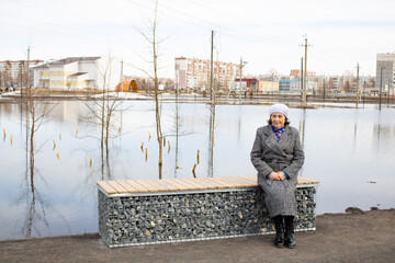 A lonely elderly woman is sitting on a bench. Floods in the city. The water spilled. Spring. The city was flooded with water. Grandmother next to the lake.