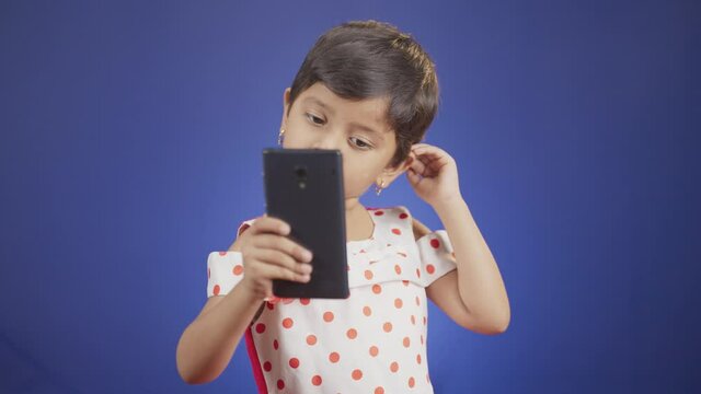 Little Girl kid or toddler taking selfie and checking photos from mobile phone on blue background - concept of technology, internet and modern lifestyle.