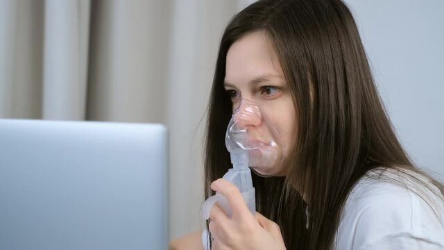 Sick young woman is using nebulizer and inhaler for treatment at home and working on laptop. She is breathing in mask. Fibrosis, covid-19, asthma, copd. Treatment, cure and procedure, therapy.
