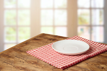 Empty white plate on wooden table with red checked tablecloth over  blurred window background. ...