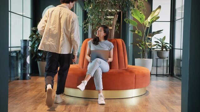 Casually dressed young man greets woman working in modern open plan office sitting on orange circular sofa in slow motion