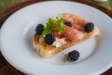 Snack, open sandwich with prosciutto, cream cheese and fresh blackberries on a white plate on a green concrete background. Sandwich recipes.