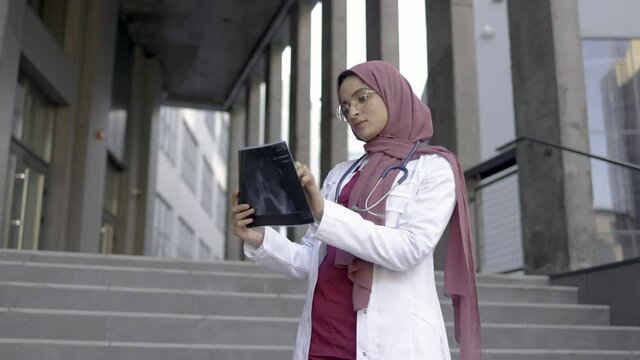 Young focused skilled pretty Arabian Muslim female doctor in hijab and medical uniform, standing outside of modern clinic at stairs, and pensively analyzing patient's skull x-ray scan