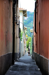 A narrow passage between houses with bright orange walls in a southern European city. A woman in a black dress is standing on the path.