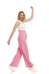Naklejka premium Happy Young Woman In Striped Loose Pants Is Walking With Arm Raised. Side View.