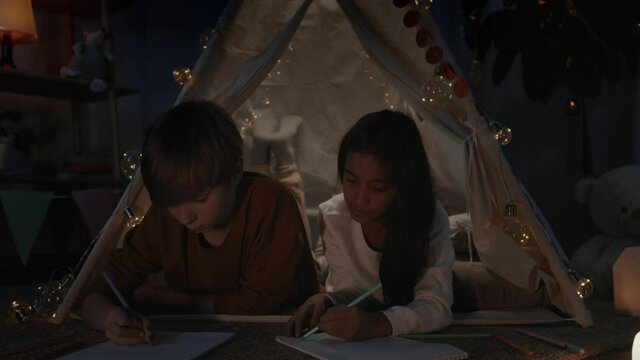 Young diverse boy and girl drawing with colored pencils while spending free time in evening. Teenagers lying on floor and talking in decorative tent while creating pictures.