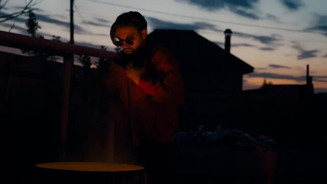 A European man in glasses and a red jacket basking near a burning garbage can on his face.