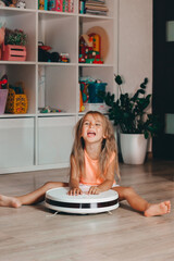 A little girl sits in the children's room on the floor with a robot vacuum cleaner and smiles cheerfully.