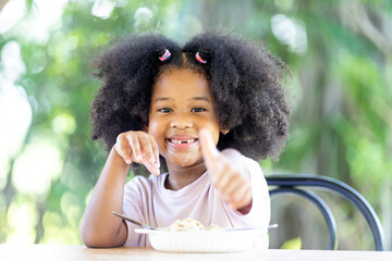 A little curly-haired African American girl sits at the table eating delicious Spaghetti Carbonara. Fun, cheerful. Appetite. Childhood and eating concepts.