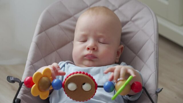 Cute 3 month old baby boy playing with toys in bouncer for kids, infant activity, child playing with wooden toys in rocking chair. High quality 4k footage