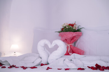 Close-up of romantic composition on bed in hotel. White towels in shape of swans. Bouquet of flowers. Rose petals.