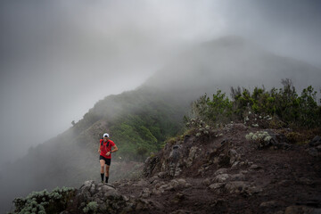 A man in a cap and red shirt doing trail running through the mountain and forest with fog and...