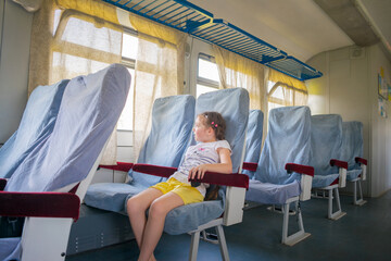 A little girl sits on herself in the train compartment. A small passenger of railway transport. The child sits in an empty electric train carriage.