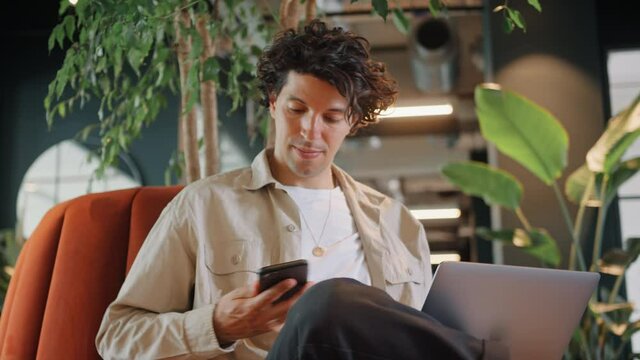 Medium shot of casually dressed young man working on laptop in modern open plan office sitting on sofa taking call on mobile phone