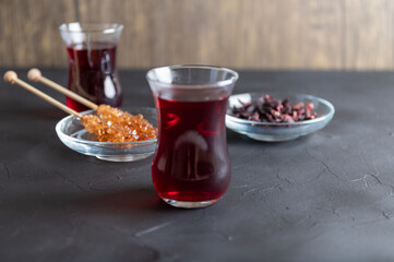Hot Karkade tea in two glasses, next to it is a glass jar with candied sugar on two wooden sticks.