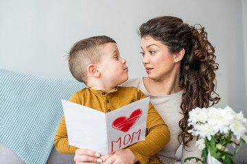 Happy little boy congratulating smiling mother and giving card with red heart during holiday celebration at home. Cheerful mother hugging son and reading handmade greeting card