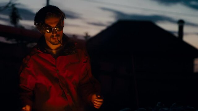 A European man in glasses and a red jacket basking near a burning garbage can on his face.