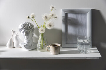 A bunch of fluffy dandelions in a chemical flask, an empty photo frame, a cup of espresso coffee, and a plaster head of David on a white chest of drawers.