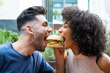 Young couple of multiethnic lovers bite the same cheeseburger, two friends having moment of fun...
