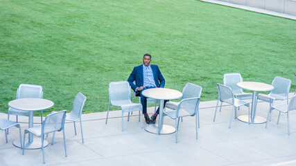 A young handsome black businessman is sitting on a chair by a green lawn and waiting for you..