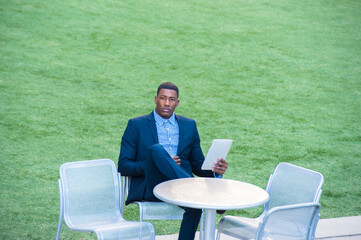 Fototapeta premium A young handsome black man is sitting on a chair by a green lawn and reading at a small computer..