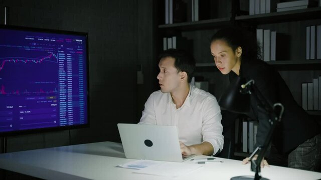 Business couple looking at monitors and discussing stock investments until nightfall.
