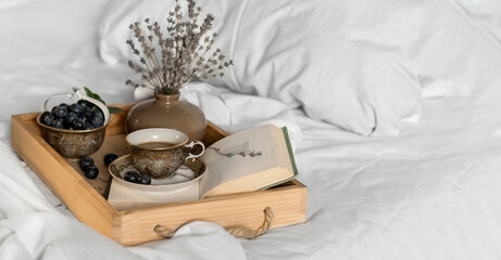 The atmosphere of a romantic morning. Cup of coffee on an opened book and blueberries on a wooden tray on a white bed. Home interior, lifestyle.