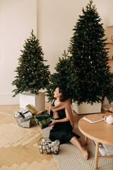 The concept of a Christmas party. A beautiful attractive young woman in an elegant dress is sitting near the Christmas tree in a decorated room in a cozy house, a selective focus