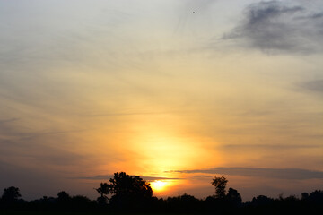 a sunset with a bright orange sun behind a silhouette of trees and a cloudy sky.